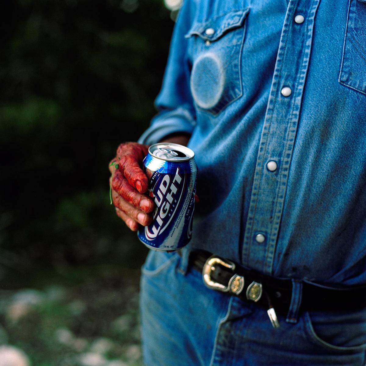 A person in a denim shirt and jeans holds a can of Bud Light with a blood-stained hand.