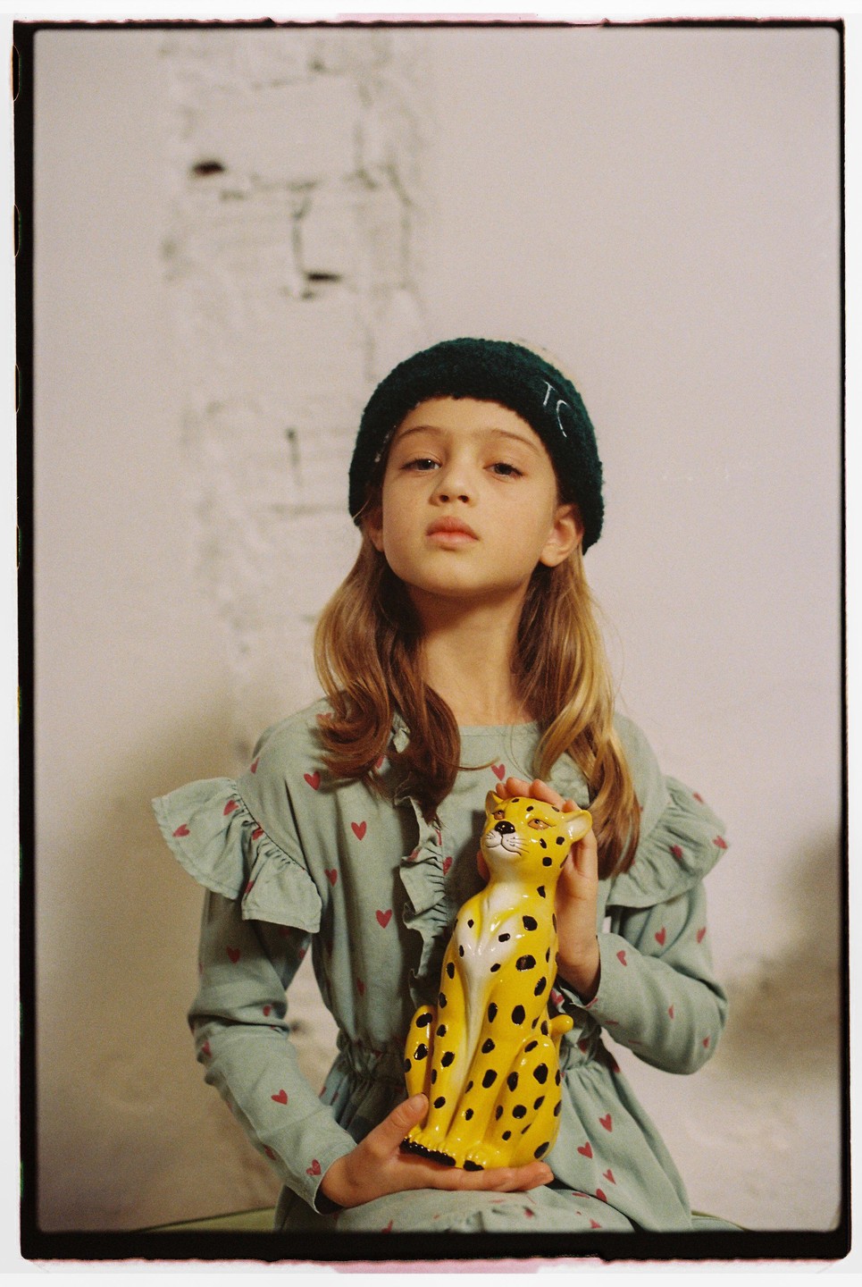 A child wearing a green beanie and a dress with red hearts holds a yellow leopard figurine.