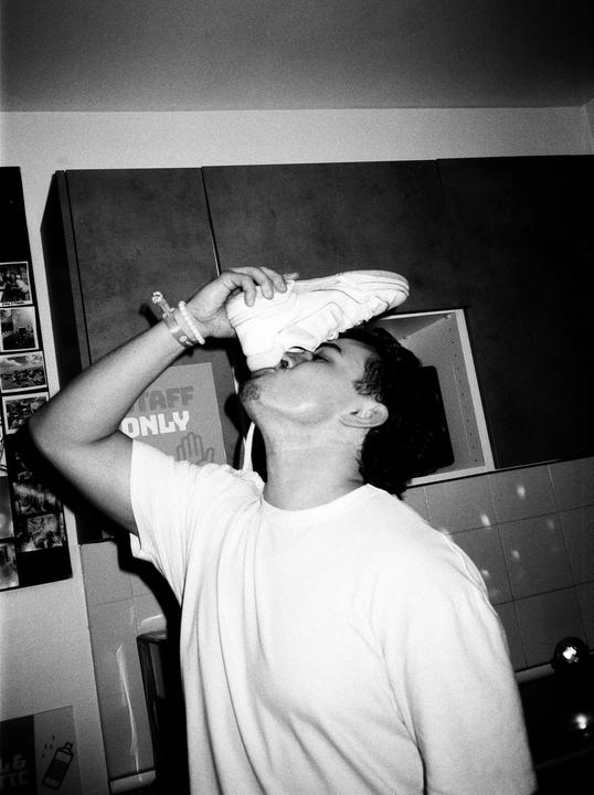 A man in a white t-shirt is drinking from a shoe in a room with cabinets and a "staff only" sign.