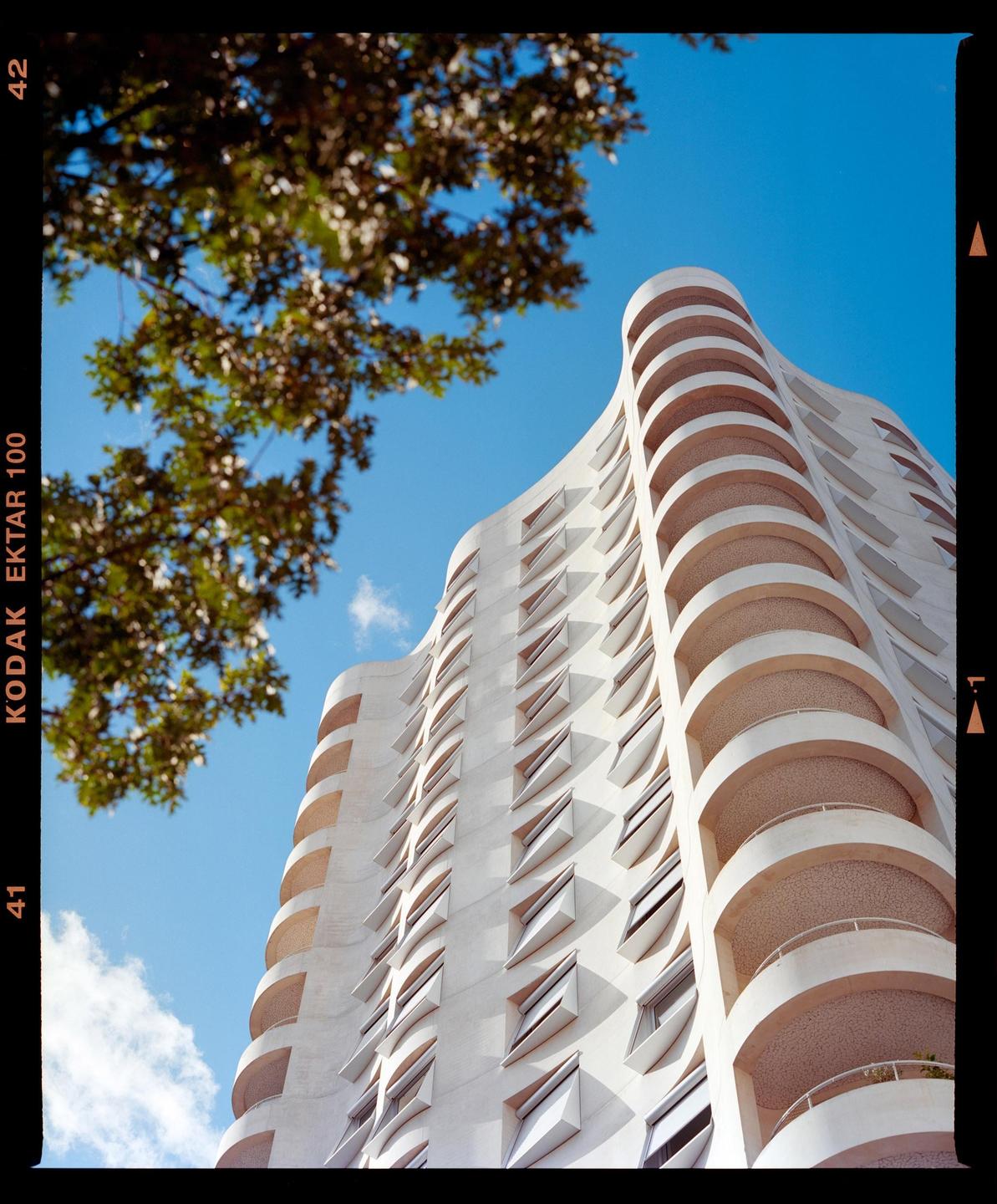 A tall, wavy white building with many windows and balconies stands against a clear blue sky, with green tree branches in the upper left corner.