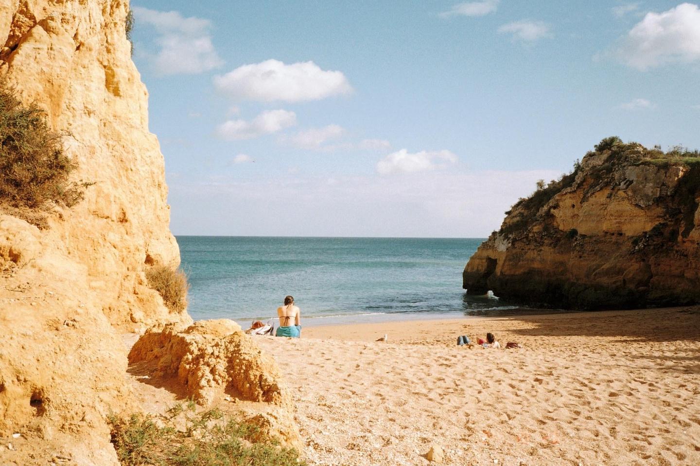 A woman sits on a sandy beach between two large rock formations, looking out at the ocean.