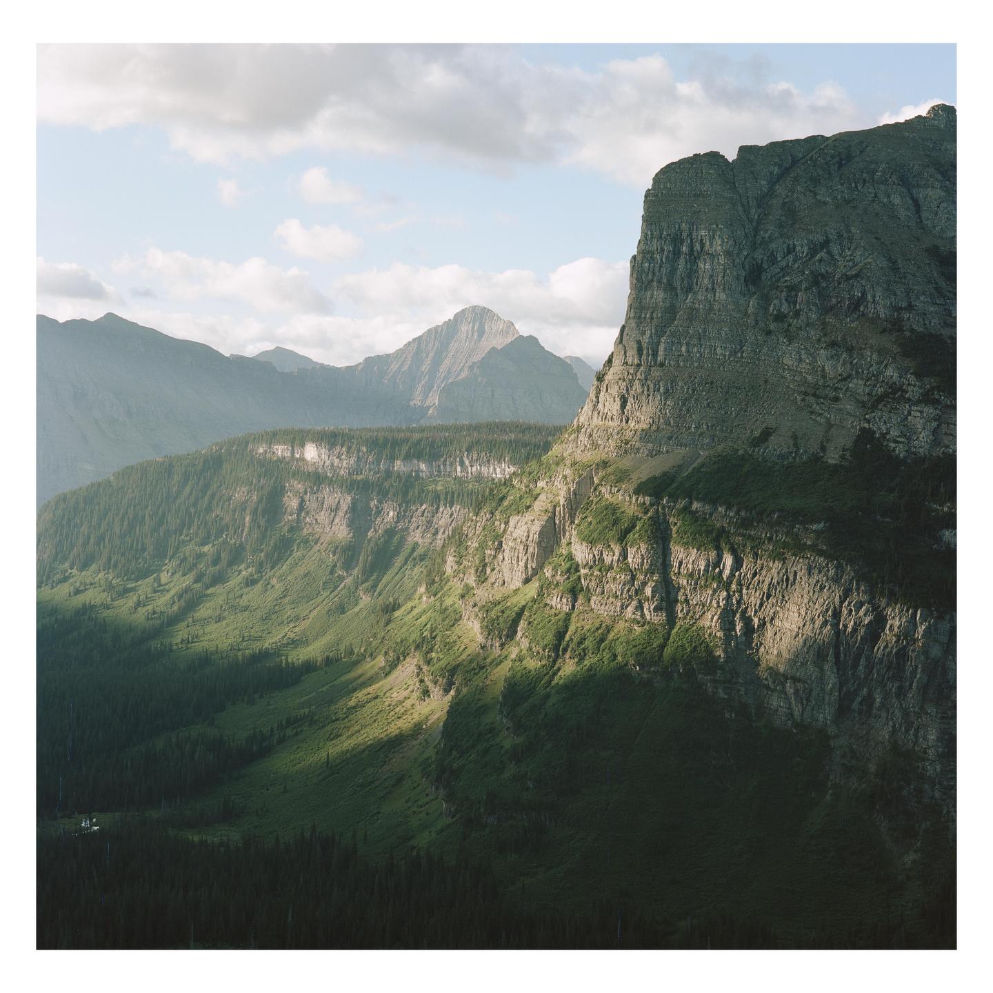 A large, rocky mountain cliff is illuminated by a patch of sunlight, with a valley of trees and other mountains in the background.