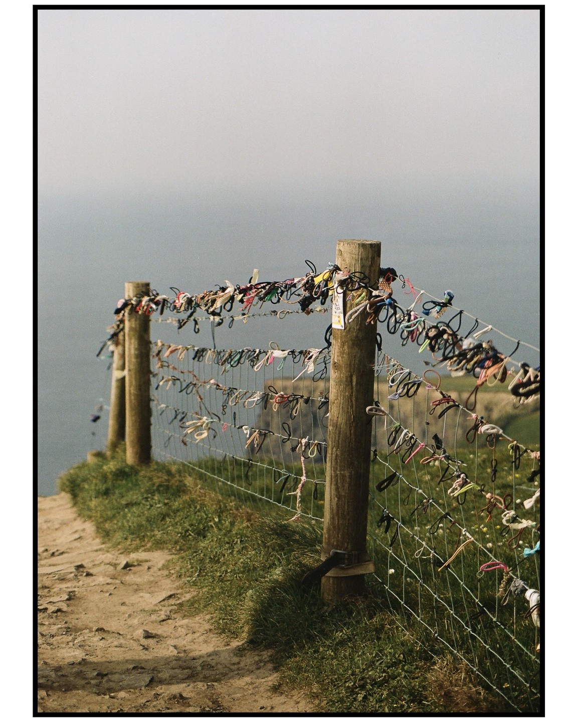 A fence adorned with colorful hair ties stands on a cliff overlooking the ocean.