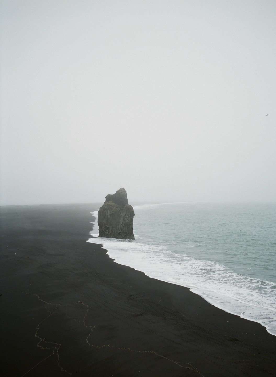 A large rock formation stands in the ocean next to a black sand beach under a hazy sky.