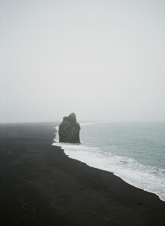 A large rock formation stands in the ocean next to a black sand beach under a hazy sky.