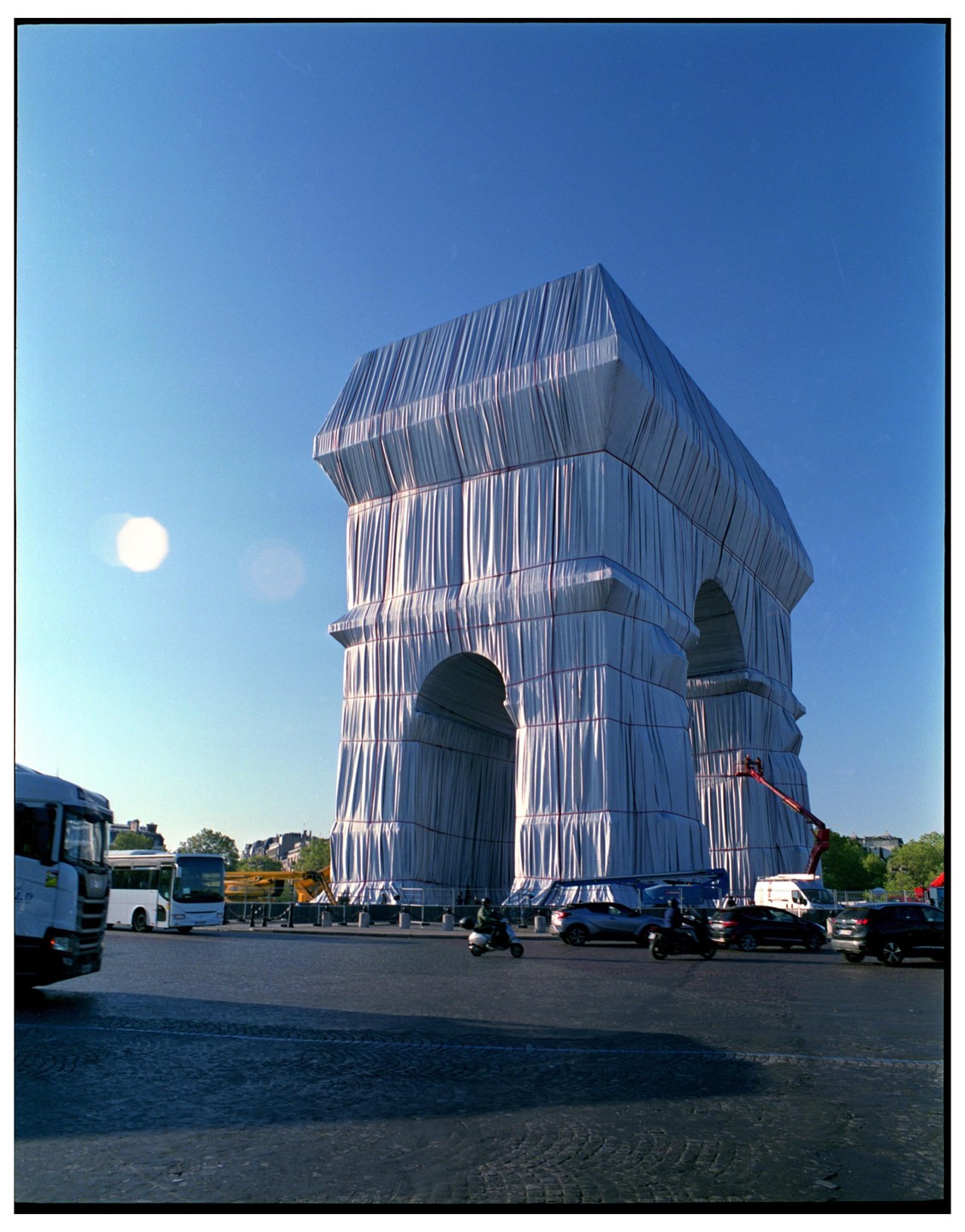 The Arc de Triomphe is wrapped in white fabric with red ropes under a clear blue sky, with vehicles and people on the surrounding street.