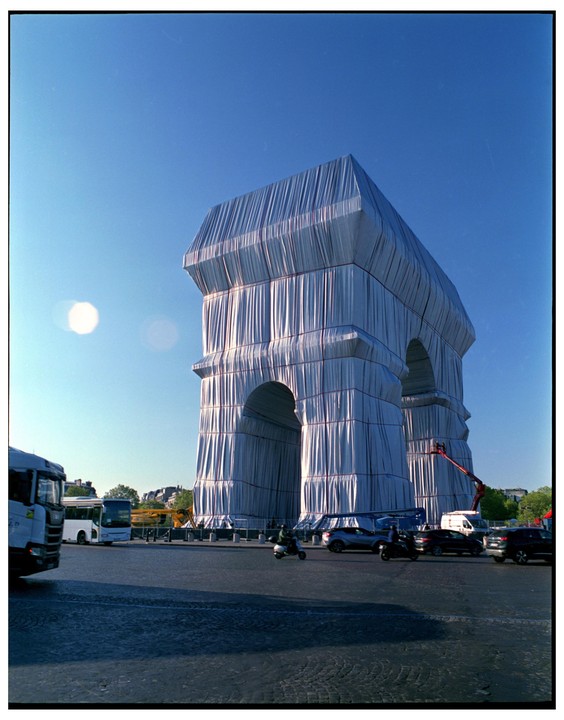 The Arc de Triomphe is wrapped in white fabric with red ropes under a clear blue sky, with vehicles and people on the surrounding street.