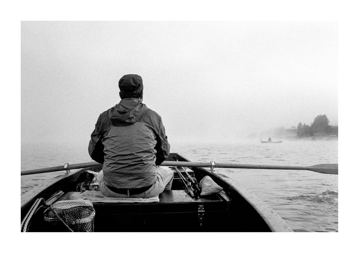 A man sits in a rowboat on a foggy lake, with another boat visible in the distance.