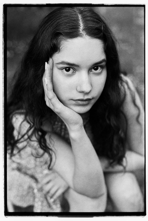 A young woman with dark, wavy hair rests her chin on her hand while looking directly at the viewer.