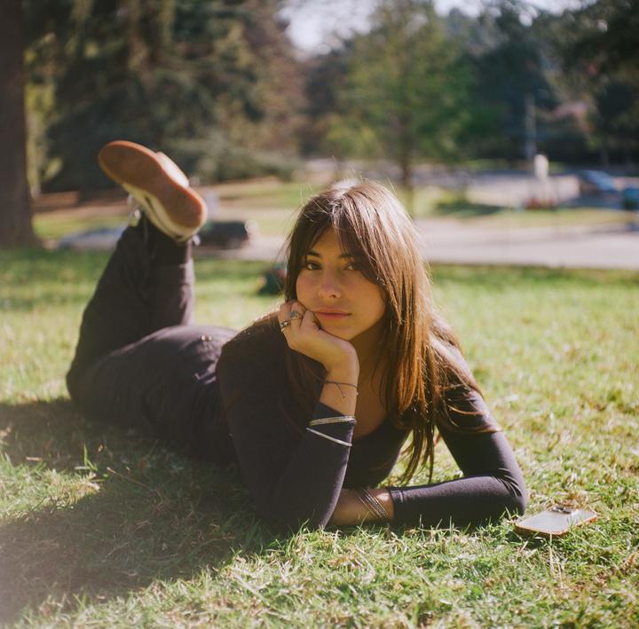 A woman with long hair is lying on her stomach in the grass, resting her chin on her hand.