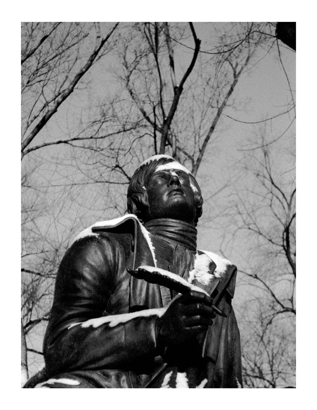A statue of a man holding a quill is covered in snow, with bare tree branches in the background.