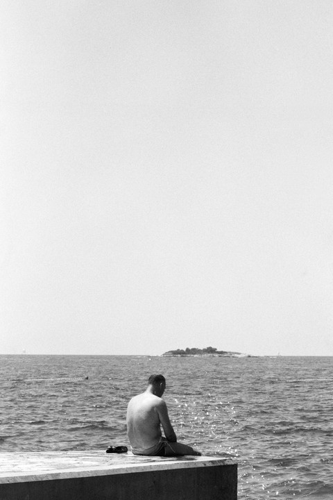 A man sits on a concrete ledge looking out at the ocean with an island in the distance.