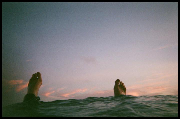 Two feet emerge from the water against a pastel sky with hints of pink clouds.
