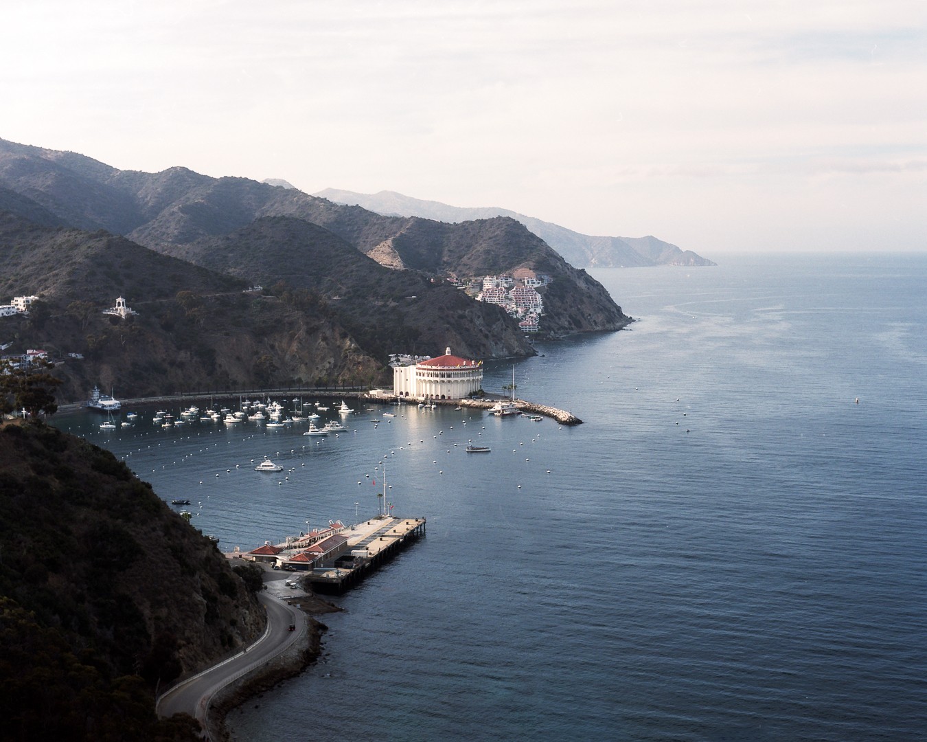 A wide view shows a bay with many boats, a large circular building with a red roof, a long pier, and a winding road along the coast, all surrounded by hills.