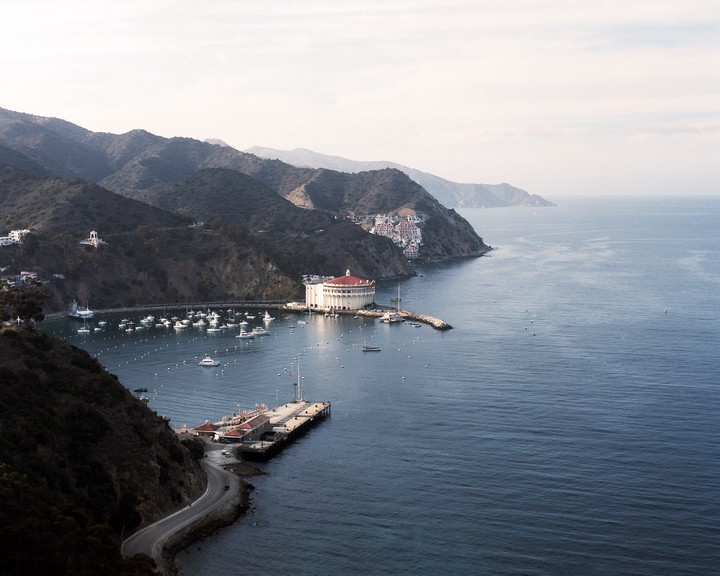A wide view shows a bay with many boats, a large circular building with a red roof, a long pier, and a winding road along the coast, all surrounded by hills.