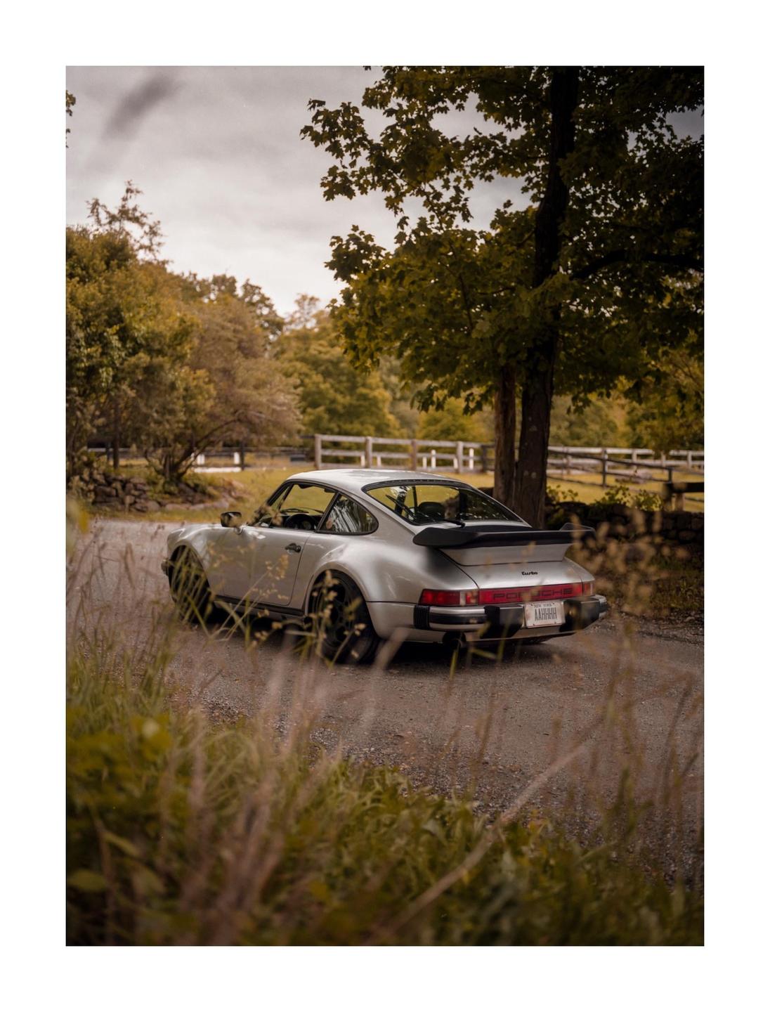 A silver car is parked on a gravel road surrounded by trees and tall grass.