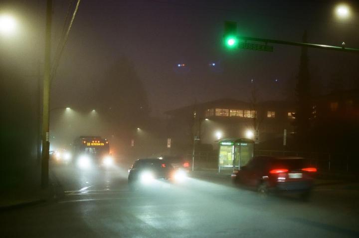 A foggy street at night shows cars with bright headlights and a green traffic light.