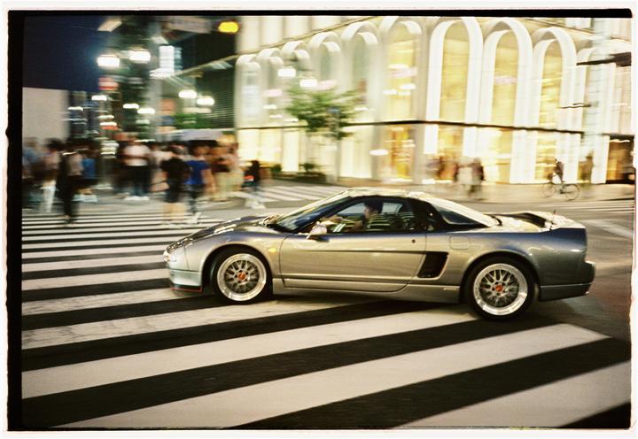 A silver sports car drives across a crosswalk at night with blurred people and buildings in the background.