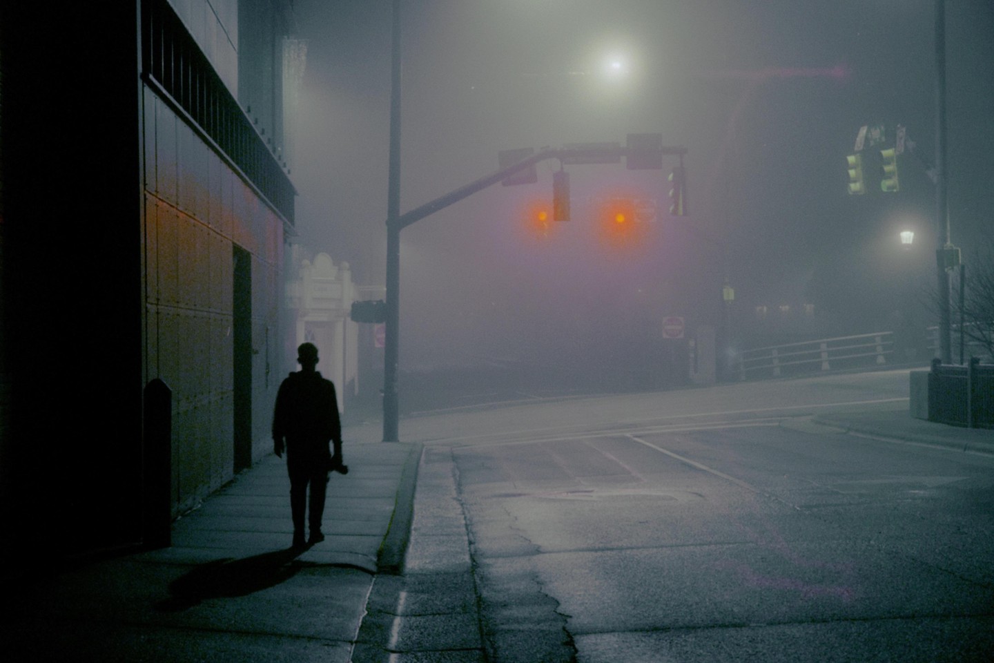 A person walks down a foggy street at night, with traffic lights glowing red in the distance.