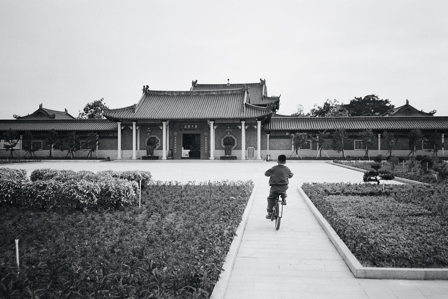 A boy rides a bicycle on a path leading to a traditional building with a tiled roof.