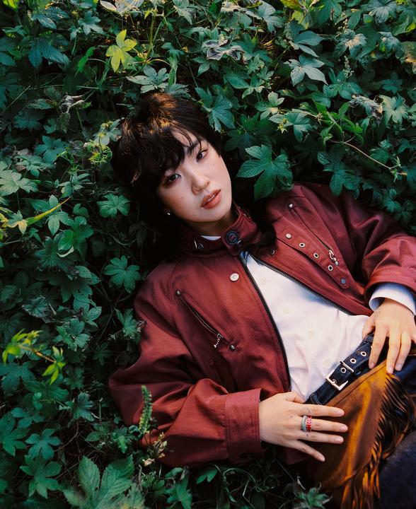 A woman with dark hair and a red jacket is lying in green foliage.