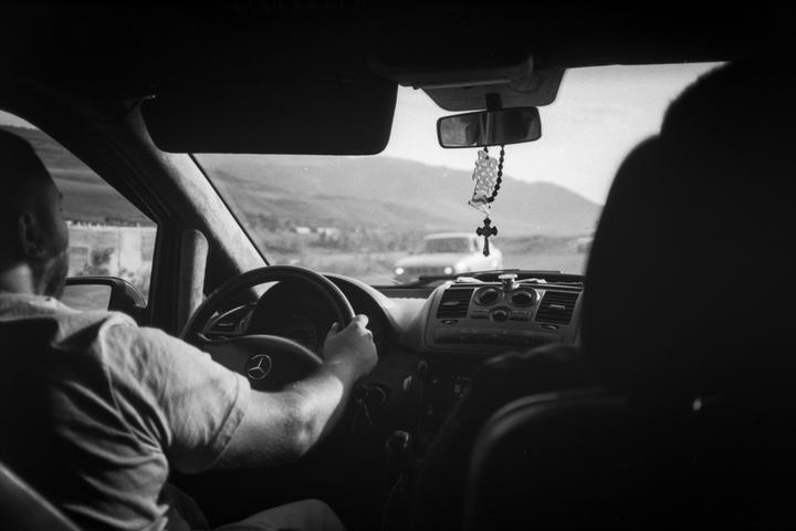A man is driving a car with a rosary hanging from the rearview mirror, with mountains in the distance.