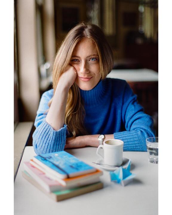 A woman in a blue sweater sits at a table with books, a coffee cup, and a glass of water.