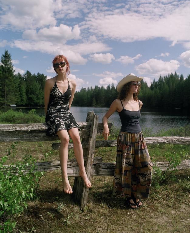 Two women are posing by a wooden fence next to a lake with a forest in the background.