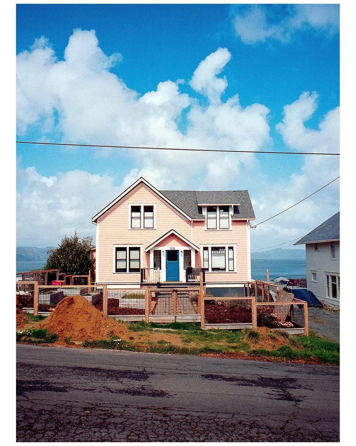 A pink house with a blue door sits on a hill overlooking a body of water under a bright blue sky with white clouds.
