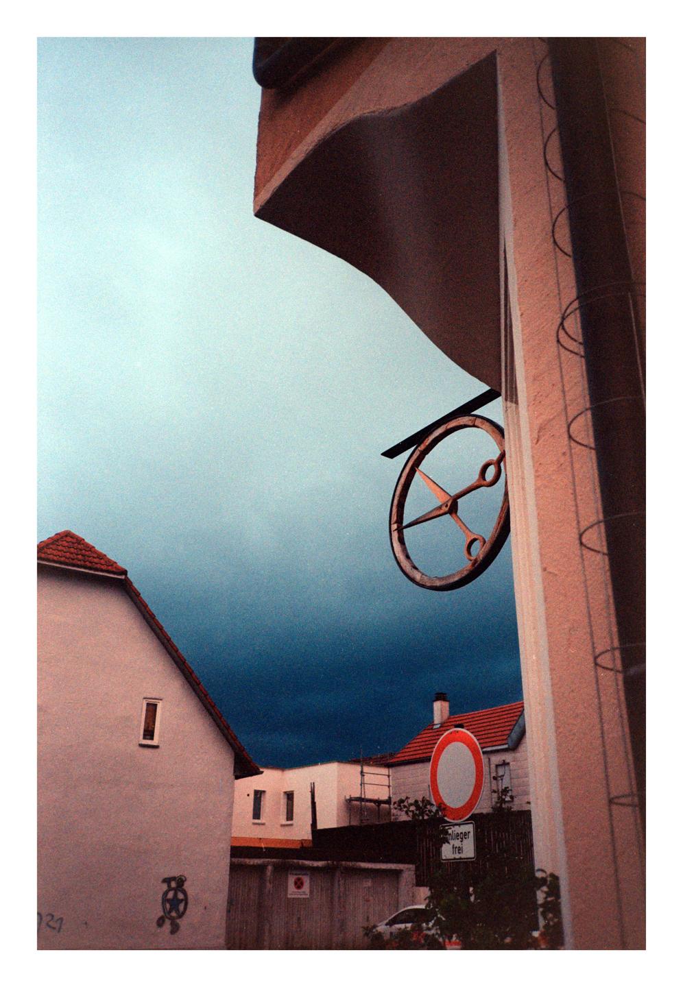 A metal sign shaped like a pair of scissors hangs from a building against a dramatic sky with dark clouds.