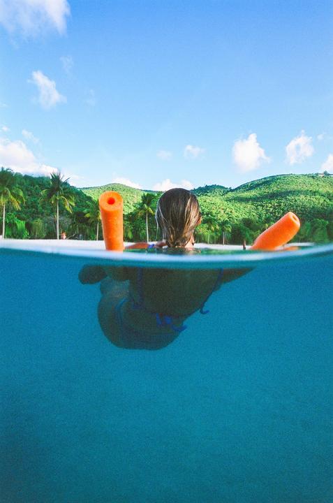 A woman floats in the ocean with two orange pool noodles, looking towards a tropical beach with palm trees and green hills.