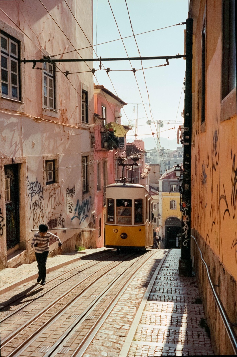 A boy runs alongside a yellow tram on a cobbled street lined with buildings covered in graffiti.