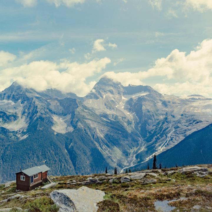 A small wooden cabin with a green roof sits on a rocky, grassy hill overlooking a valley with large mountains in the background.