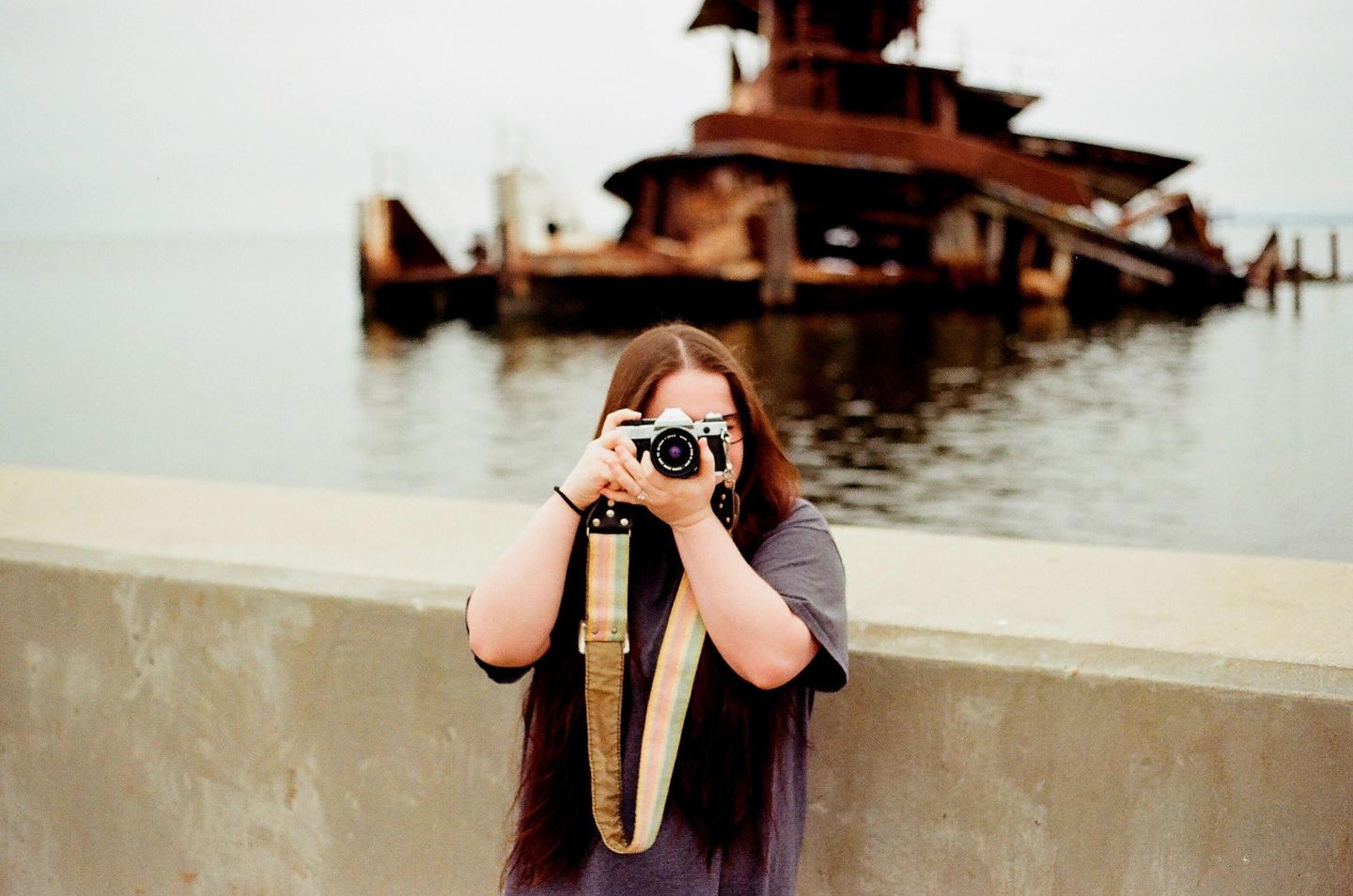 A woman with long hair holds a camera to her face, with a rusted shipwreck in the water behind her.