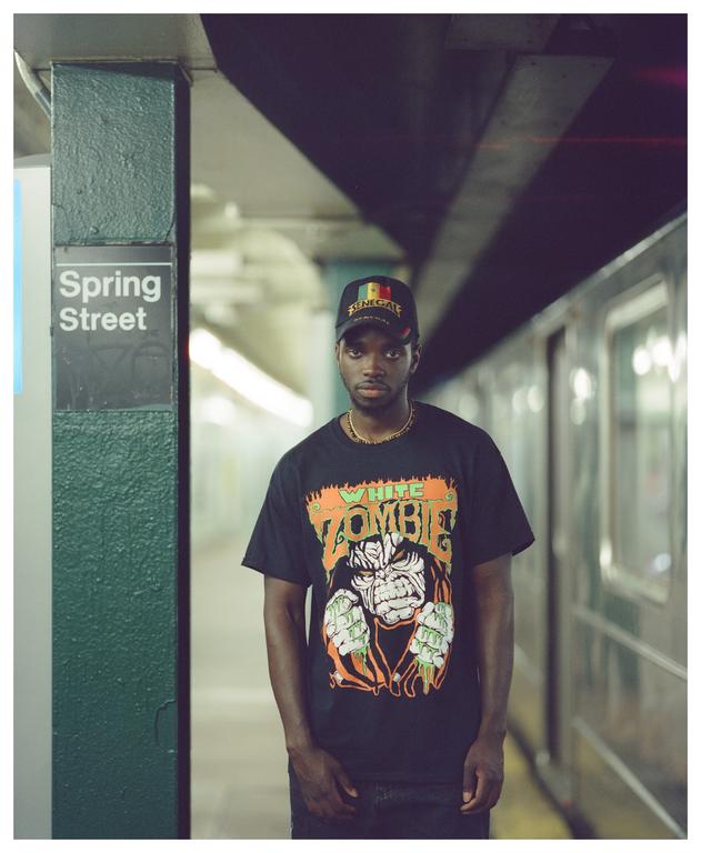 A man wearing a black t-shirt and a cap stands on a subway platform next to a sign that reads "Spring Street."