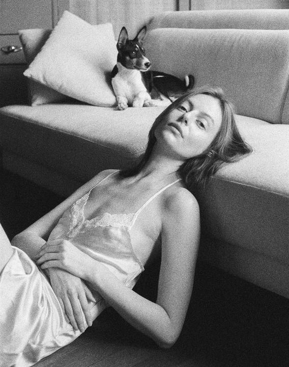 A woman in a slip lies on the floor with a dog on the couch behind her.