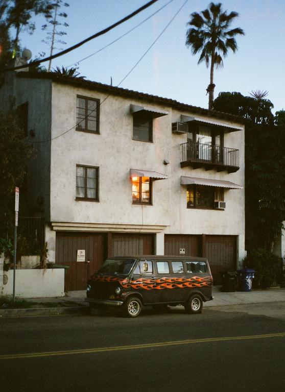 A black van with orange flames painted on its side is parked on a street in front of a white building with a palm tree in the background.