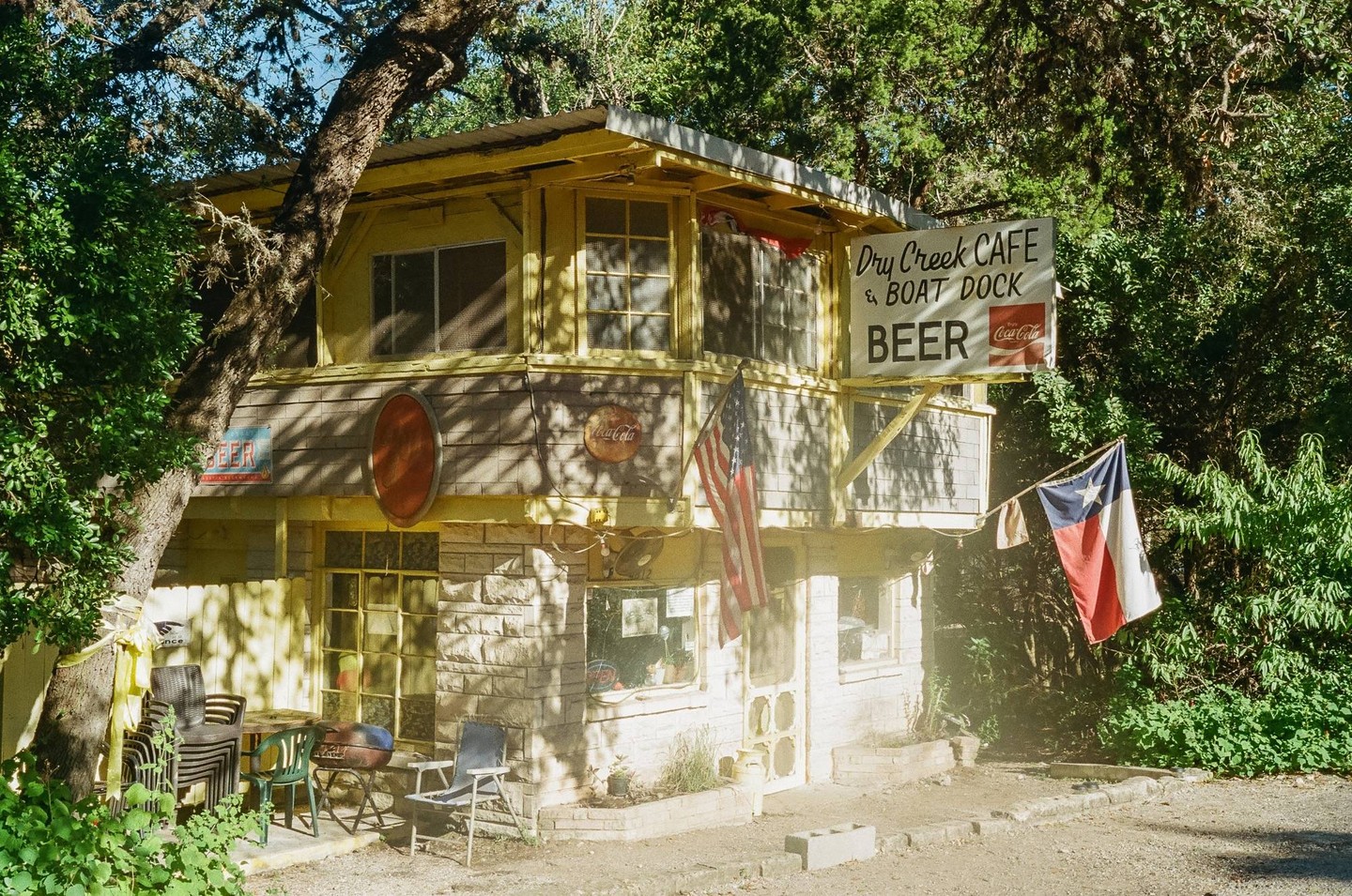 A two-story building with a "Dry Creek Cafe & Boat Dock" sign and an American flag and Texas flag hanging from it is surrounded by trees.