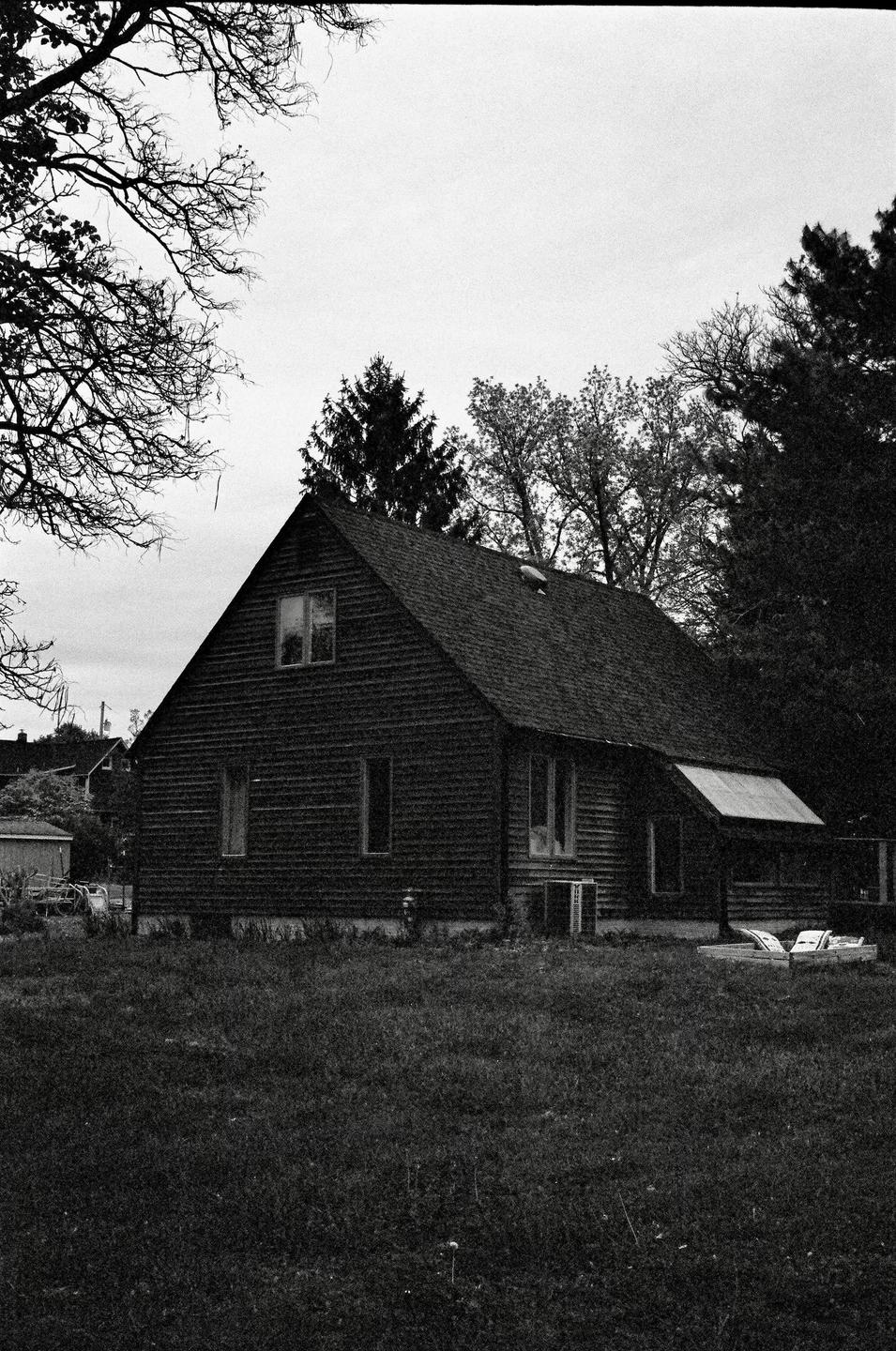 A dark wooden house with a steep roof stands in a grassy field surrounded by trees under a cloudy sky.