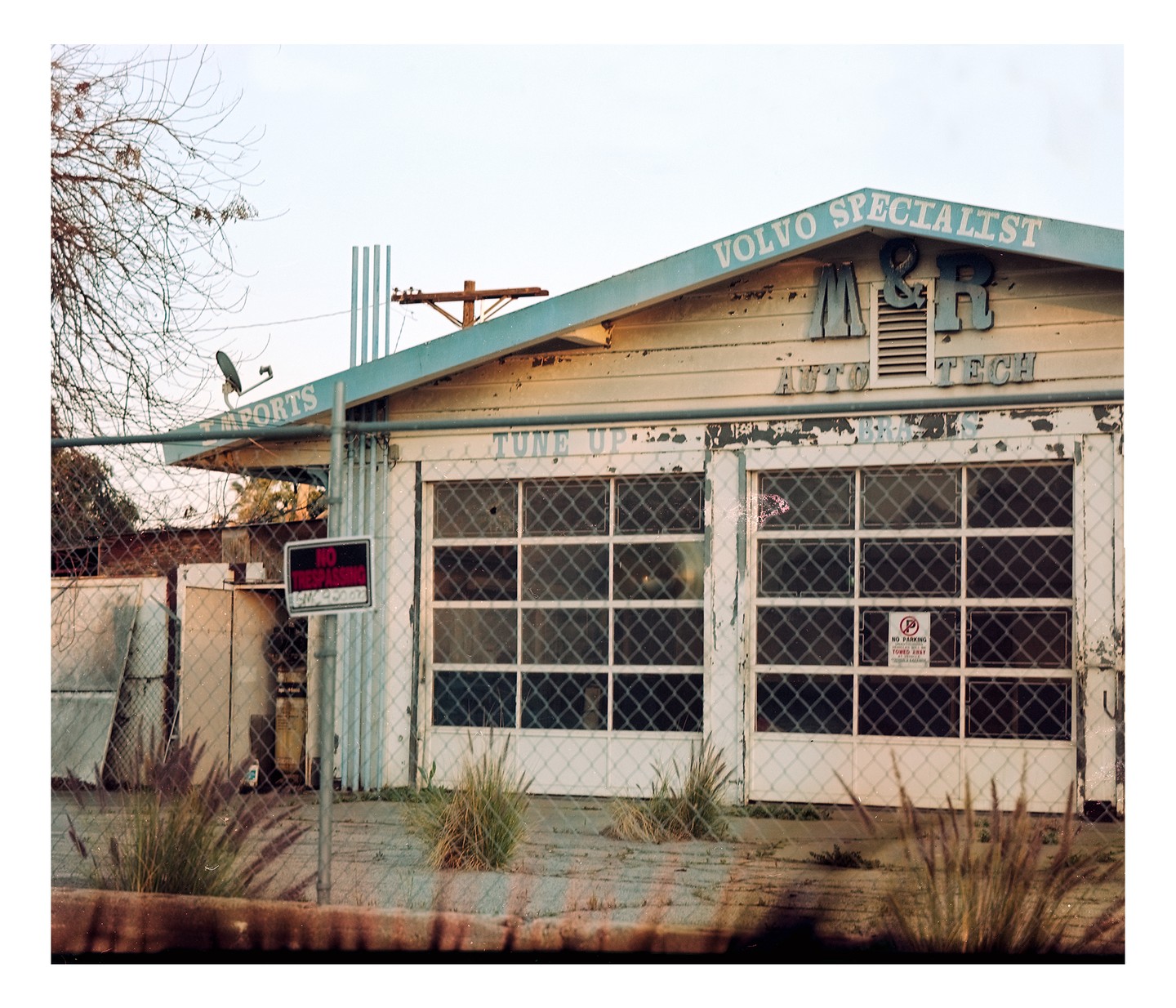 A weathered auto repair shop with a "Volvo Specialist" sign on the roof stands behind a chain-link fence.