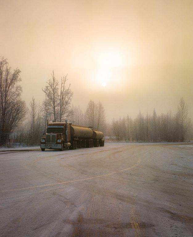 A large tanker truck is parked on a snowy road with bare trees in the background under a hazy golden sky.
