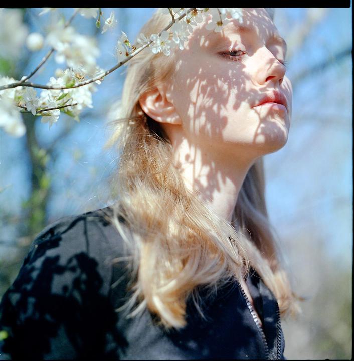 A woman with long blonde hair looks up at the sky, with shadows of leaves on her face and a branch of white blossoms in the foreground.