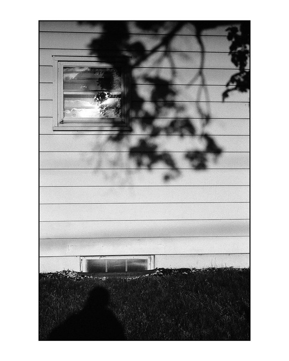 A window reflects the sky and trees on the side of a house, with a shadow of a person in the foreground.