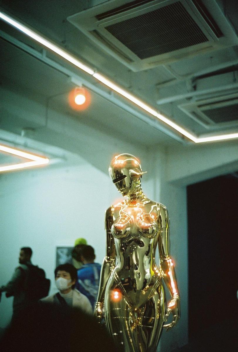 A golden robot sculpture stands in a room with people in the background.