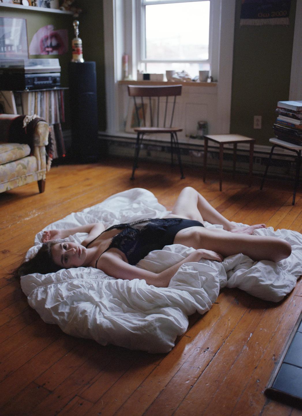 A woman in a black bodysuit is lying on a white sheet on a wooden floor in a room with a window.