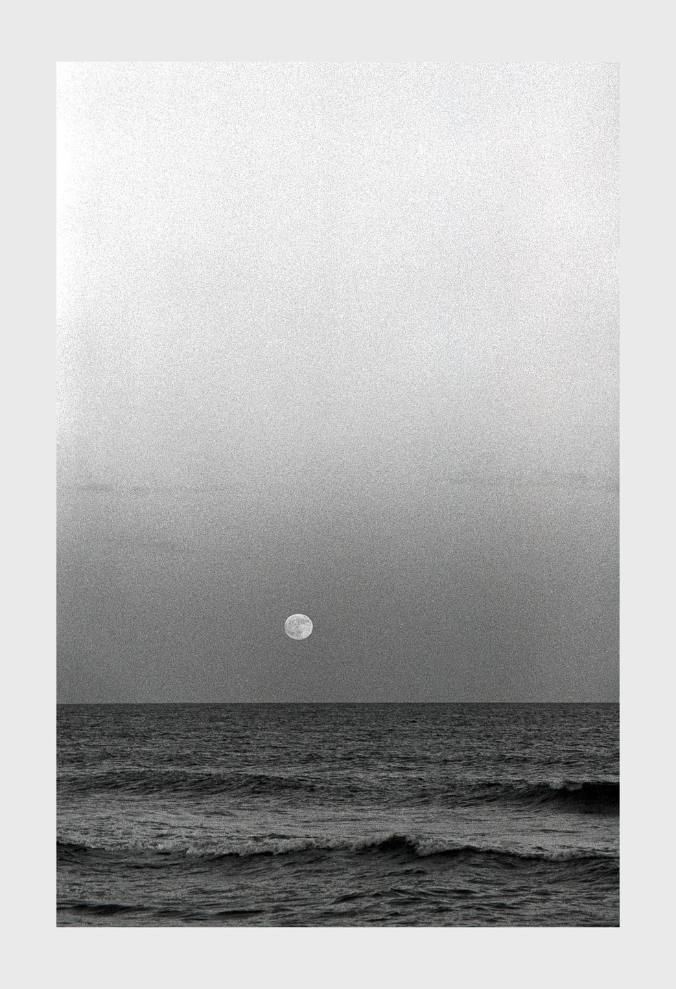 The full moon hangs over the ocean with waves crashing in the foreground.