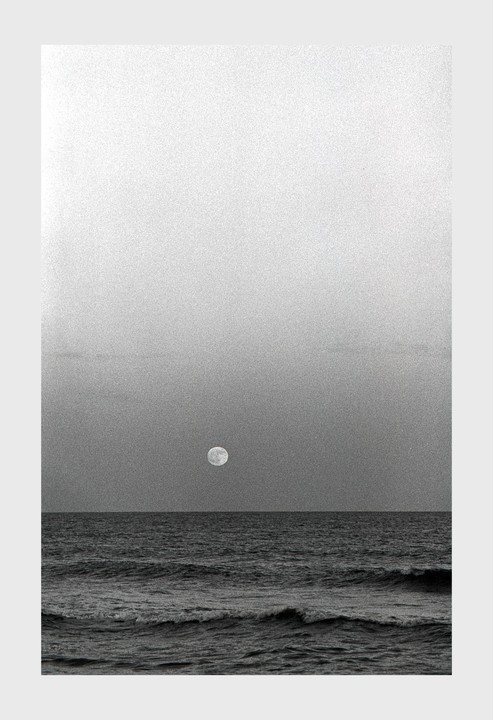 The full moon hangs over the ocean with waves crashing in the foreground.