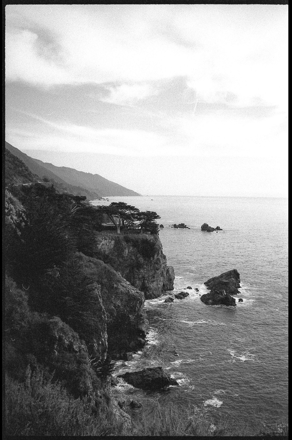 A black and white landscape shows a rugged coastline with cliffs, trees, and mountains meeting the ocean under a cloudy sky.
