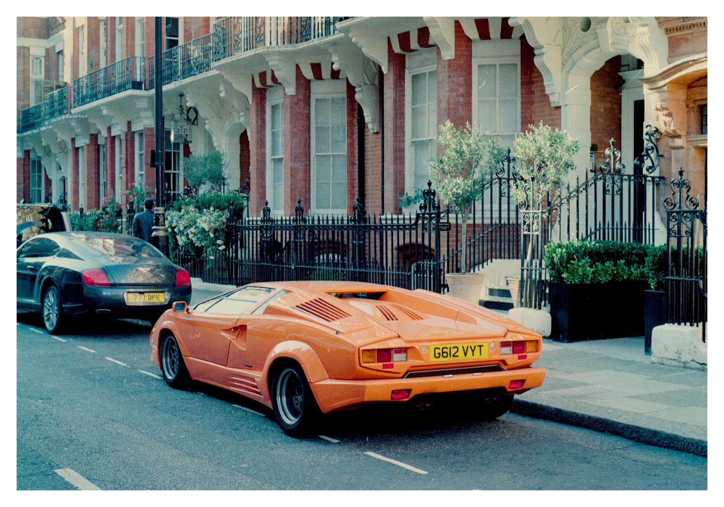 An orange sports car is parked on a street in front of a row of brick buildings with ornate iron fences.