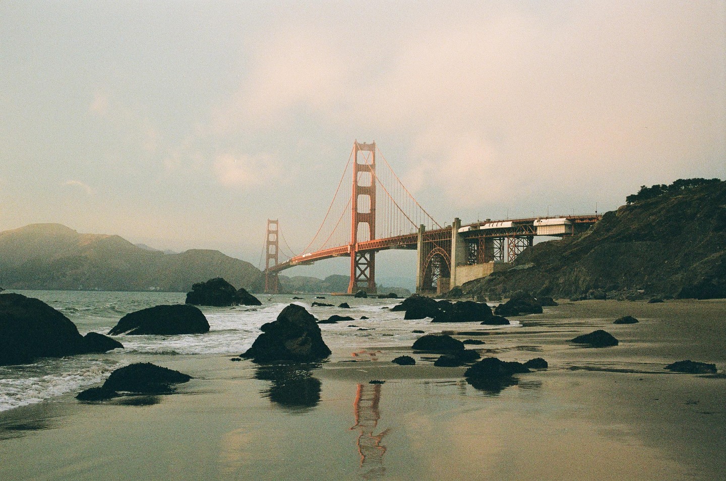 The Golden Gate Bridge is visible from a rocky beach with waves crashing on the shore.